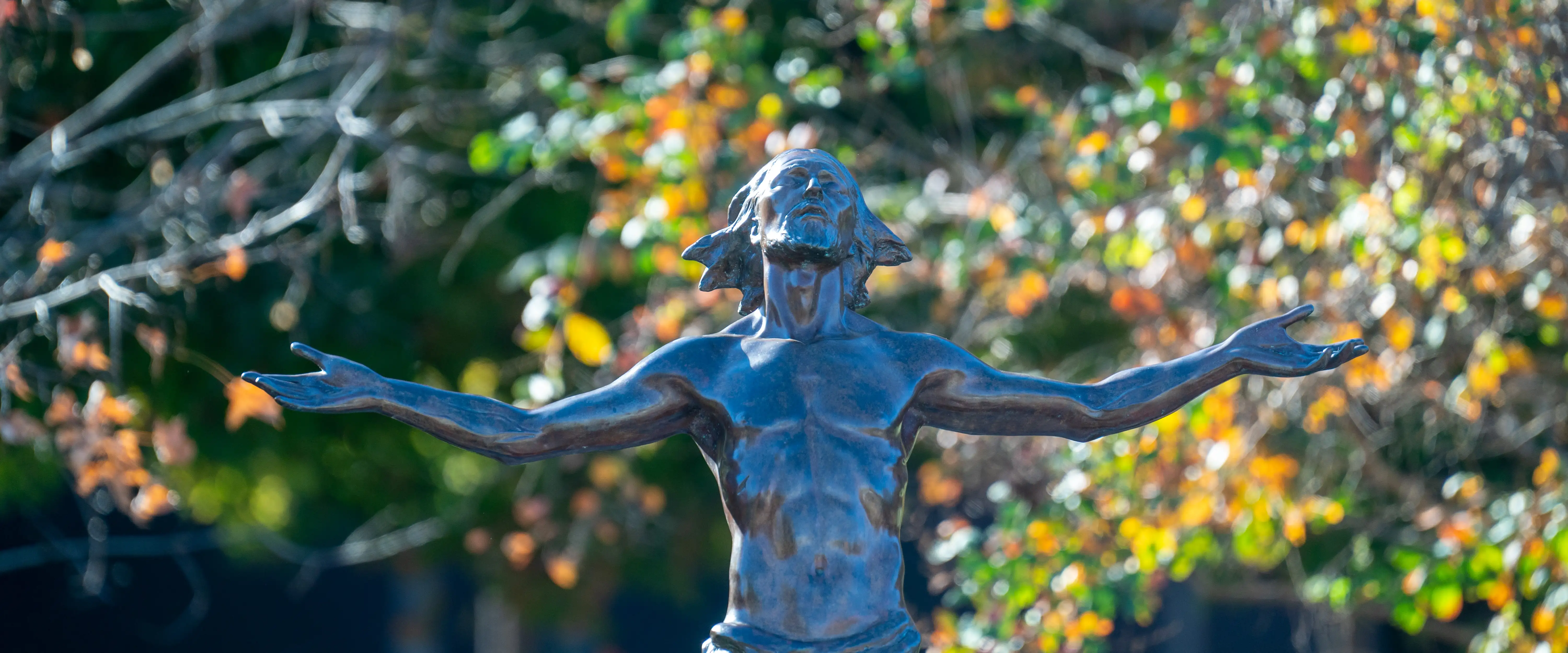 Bronze statue of Jesus with arms outstretched in front of autumn trees on Belmont’s campus.