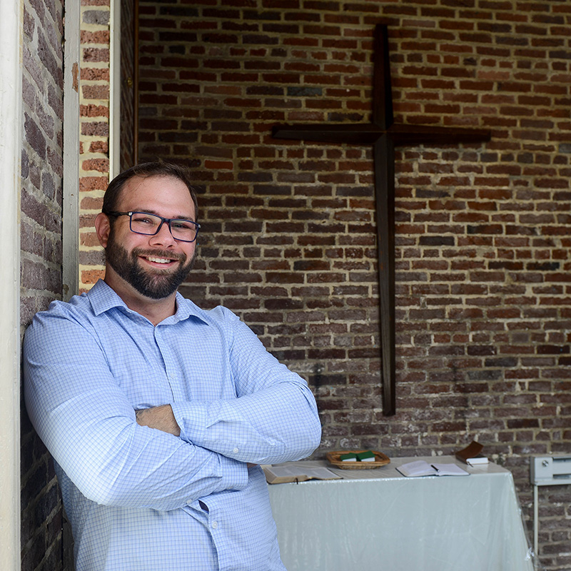 A man in glasses leans against a brick wall with a cross hanging on it.