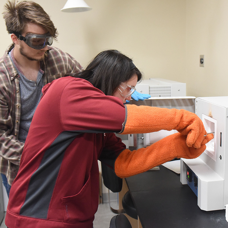 A female student wearing goggles and heat-resistant gloves uses tongs to place something into a box.