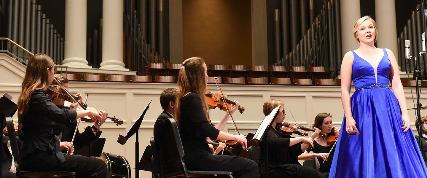 A female student stands in front of an orchestra on stage and sings.