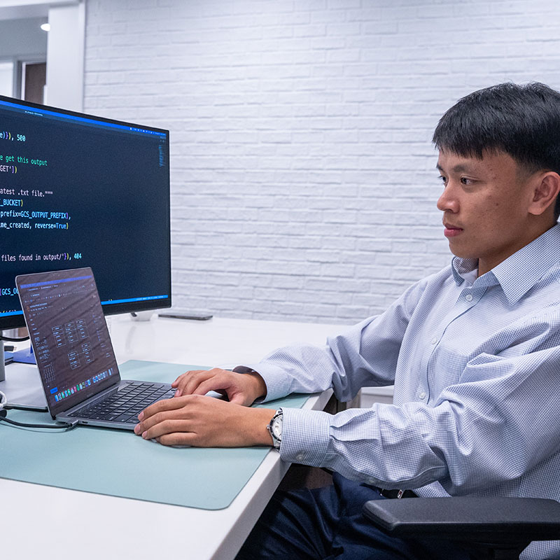 A student, dressed in a formal button-up shirt, sits at a desk working on a laptop connected to a large monitor. The screen displays lines of code, indicating that the student is engaged in a computer science or programming task. The workspace is modern and organized, with a clean, white brick wall in the background. The student's focused expression suggests deep concentration on the task at hand, highlighting a professional or academic environment dedicated to coding and problem-solving.