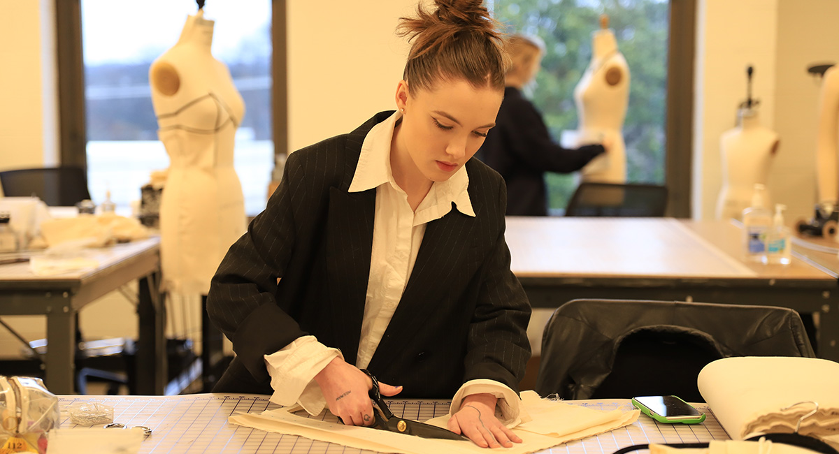 A female student stands at a drafting table and cuts a large piece of fabric with scissors.