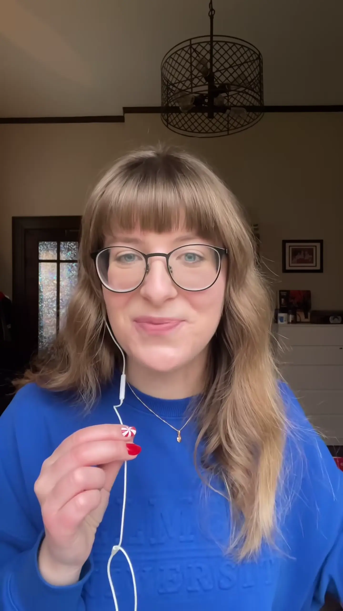 Portrait selfie of a person with shoulder-length wavy blonde hair and bangs, wearing round-framed glasses and a bright blue sweater. They are holding up one white earbud in their hand with red nail polish visible, wearing a delicate gold necklace. The background shows a warm-toned interior space with a decorative metal cage pendant light and a window.