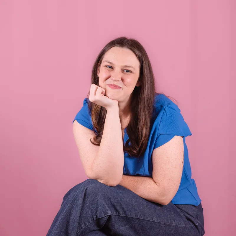 Meredith Faulkner in a blue blouse sits with her chin resting on her fist, smiling against a pink background.