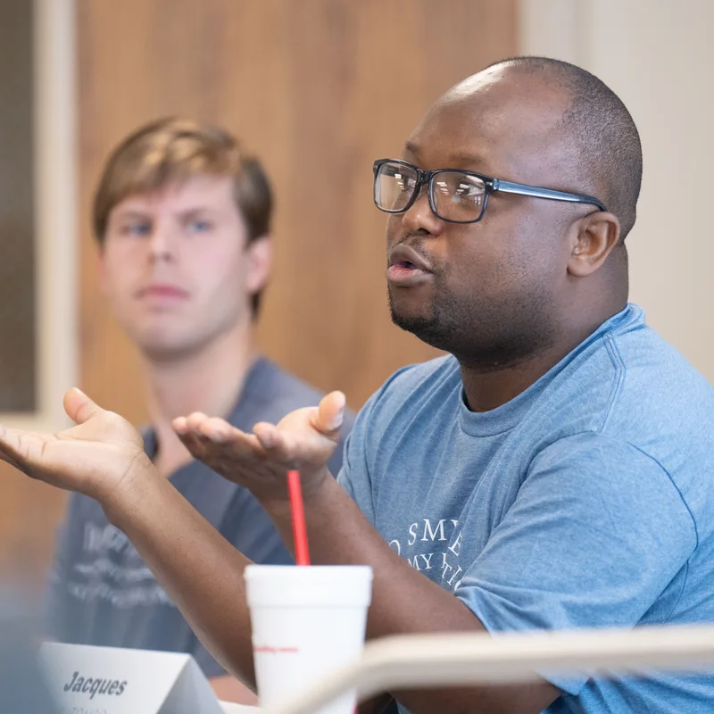 Man wearing glasses and blue shirt speaking animatedly at a panel event, with another attendee listening in the background