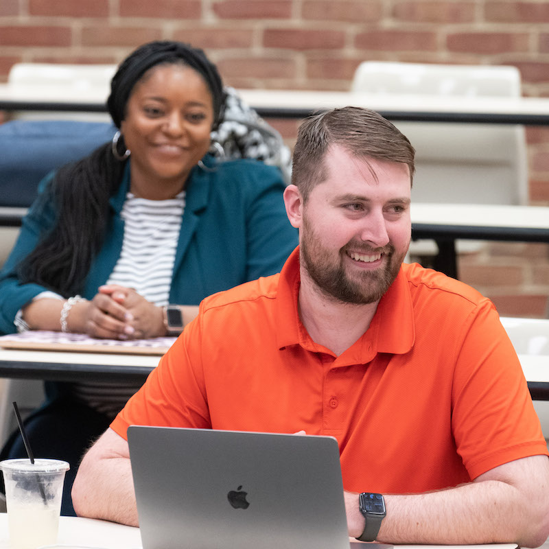 A male student in an orange polo shirt sits at a desk and works on his laptop.