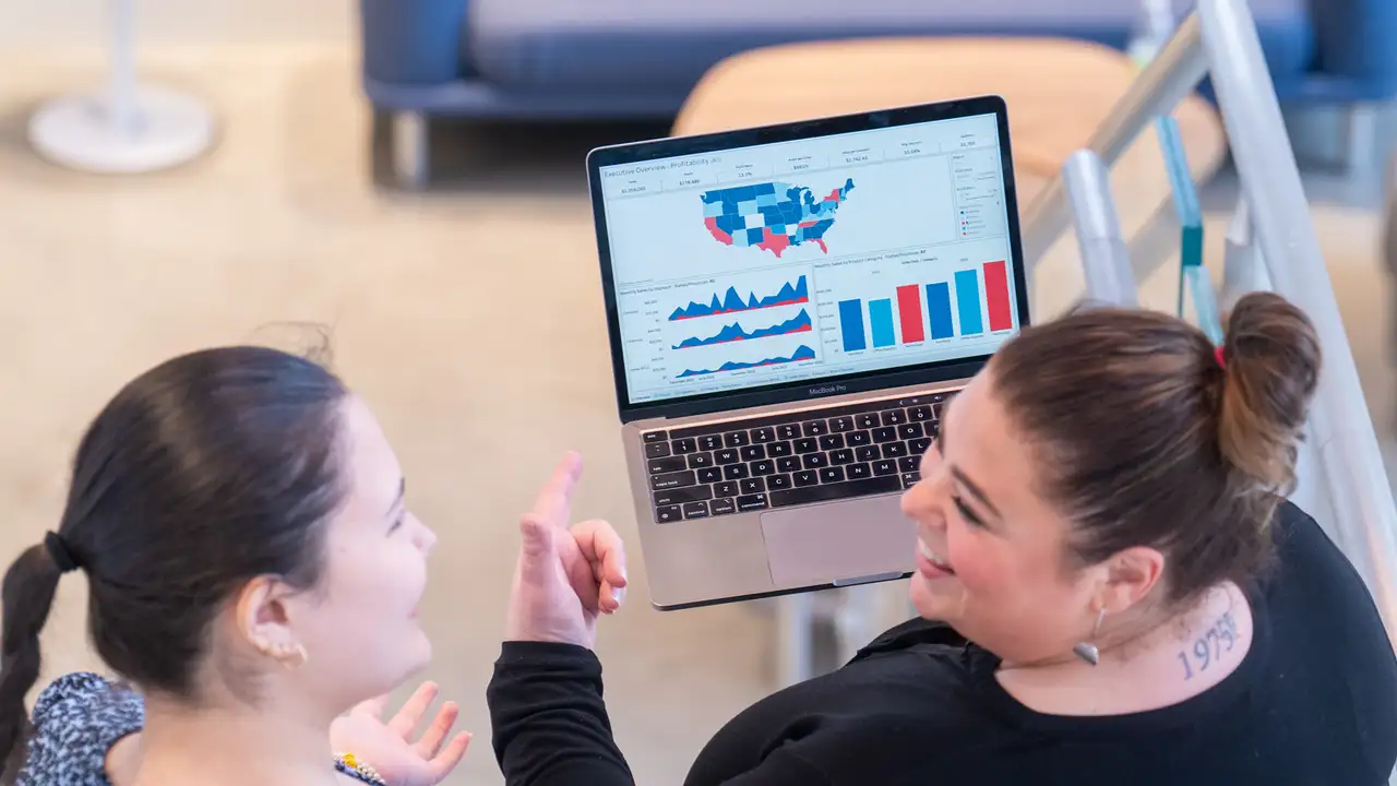 Two women engaging in conversation; one woman holding a laptop and pointing at a analytical chart