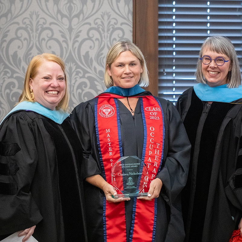 Three women in academic regalia smile for a photo as the graduate in the center holds a glass award. The graduate wears a red Master of Occupational Therapy Class of 2023 stole, while the two faculty members on either side stand beside her during the recognition ceremony.