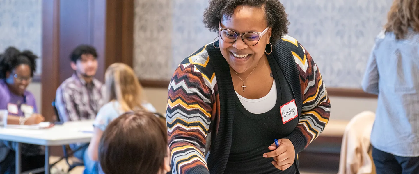 A smiling woman wearing glasses and a name tag interacts with another person during a social work event, with other attendees seated at tables in the background.