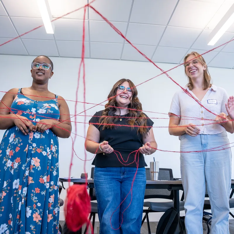 Three women participating in a team-building workshop with pink string forming a web pattern, symbolizing mental health support and community connection