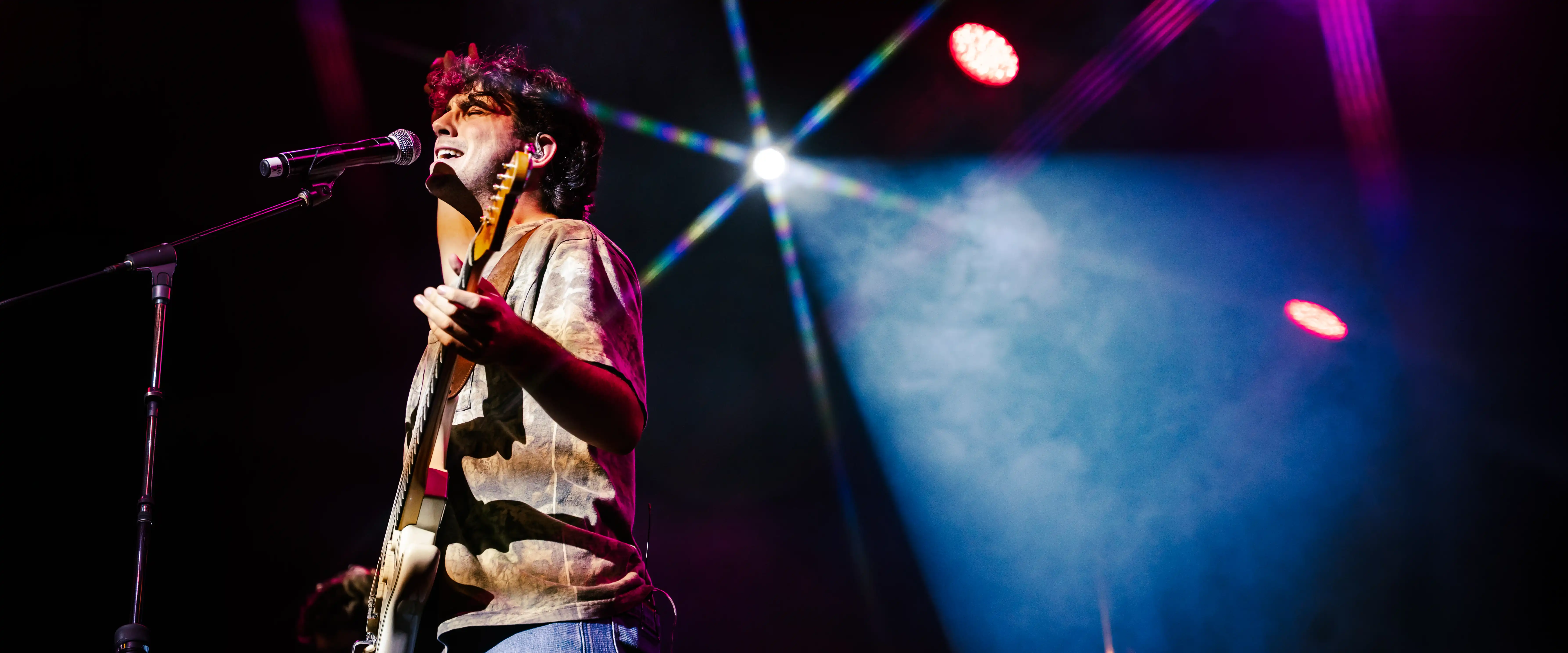 Student performs onstage, singing and playing guitar under colorful concert lighting during a live music performance.