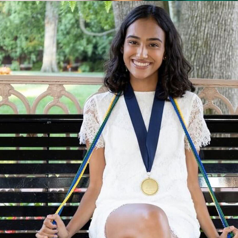 alumna Aleya Prasad sitting while wearing a white dress and graduation cords