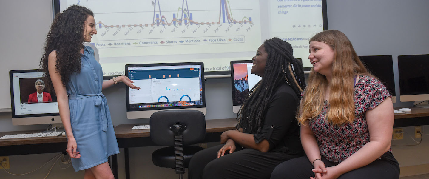 Three students stand around computers learning in a digital lab