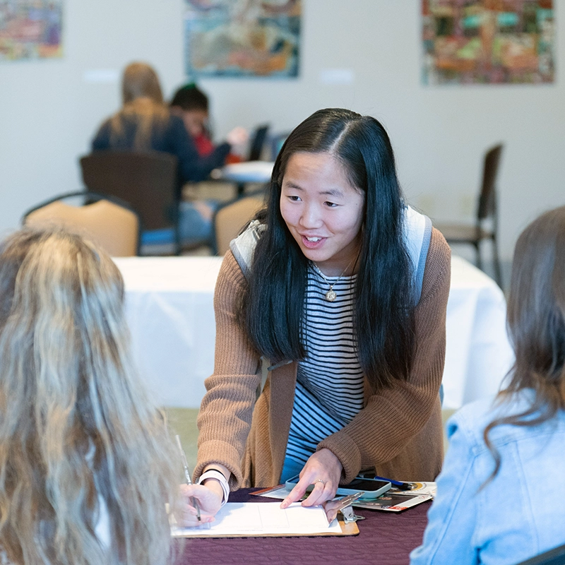An Asian woman bends over a table while conversing with two other women and writing on a clipboard.