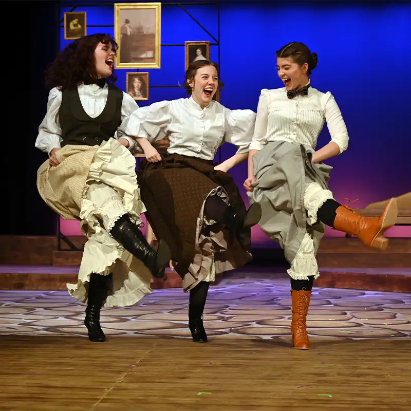 Three female performers in historical costumes joyfully dance on stage, lifting their legs in unison. They are smiling and engaged in their performance, set against a colorful backdrop with framed portraits and a vintage couch, creating an energetic and lively atmosphere.