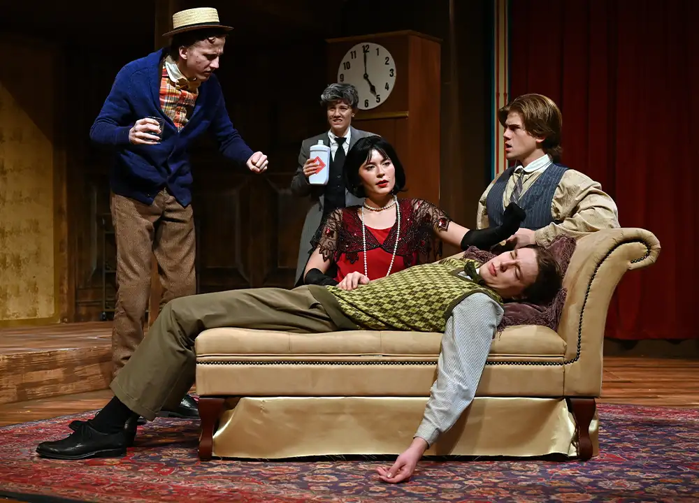 A scene from a theatrical performance featuring five actors in period costumes. One actor, lying on a couch, appears to be in distress while others surround him, engaging in animated conversation. The stage is set with a vintage aesthetic, including a clock and rich textures, creating a dramatic atmosphere for the play.