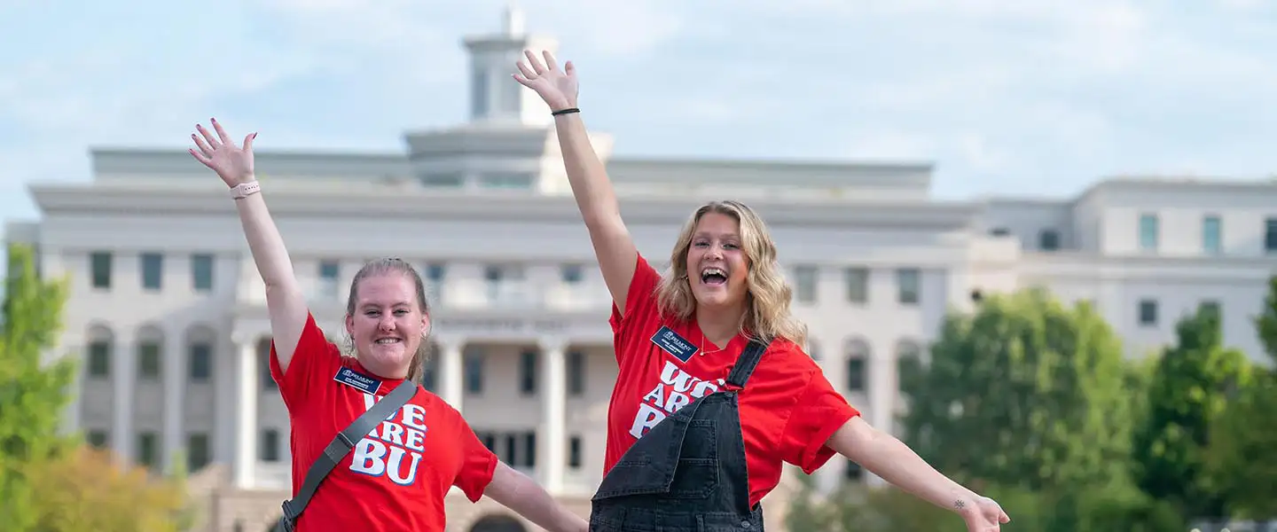 Two student volunteers smile and pose with their arms out while wearing matching t-shirts outside on the main lawn