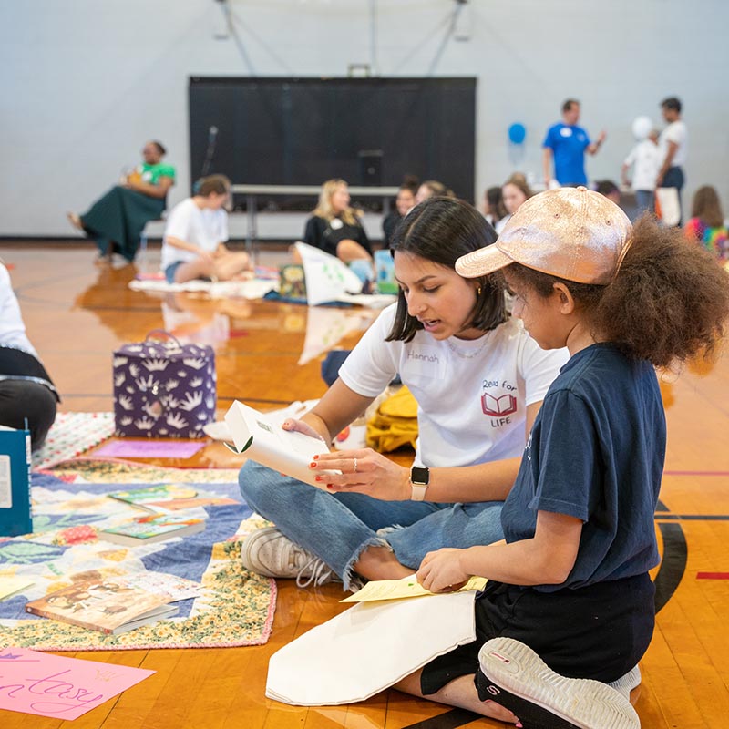 Student reading to child on the floor of a gym at family literary day 2023