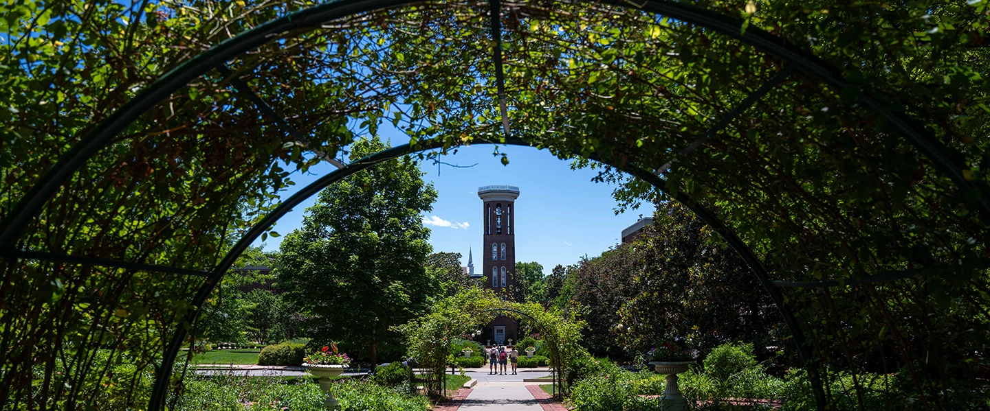 View through a leafy garden archway looking toward the bell tower with a clear blue sky in the background, surrounded by green trees and garden pathways with people walking.