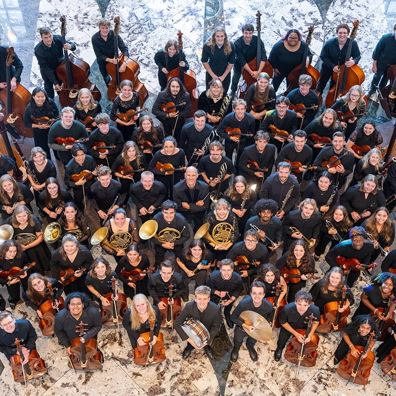 Large orchestra group portrait taken from above, with musicians holding string, brass, woodwind, and percussion instruments, arranged in rows on a marble floor.
