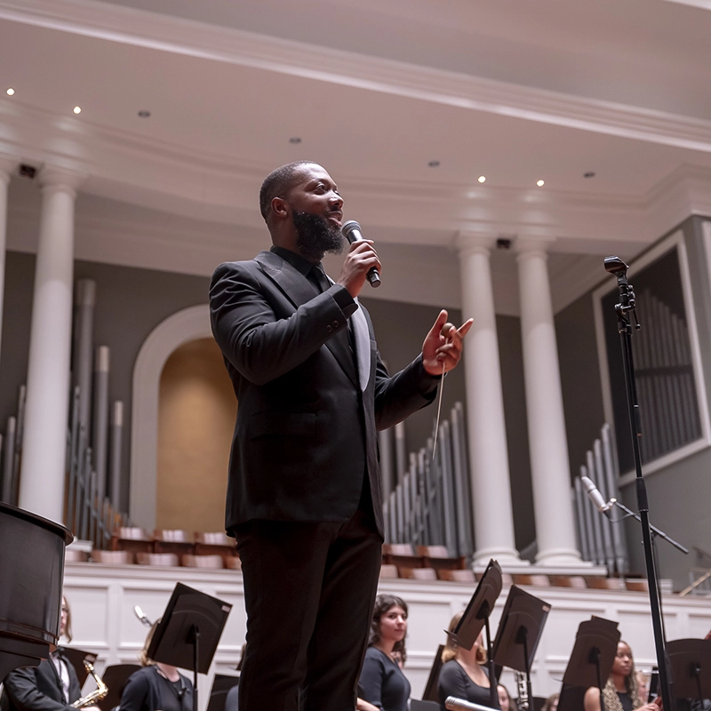 A man in a black suit speaks into a microphone on stage in a concert hall, with musicians and music stands visible behind him.