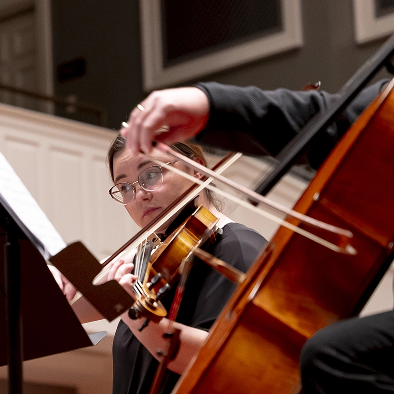 A violinist reads sheet music while playing in a performance, framed closely with a cellist’s bow and instrument in the foreground.
