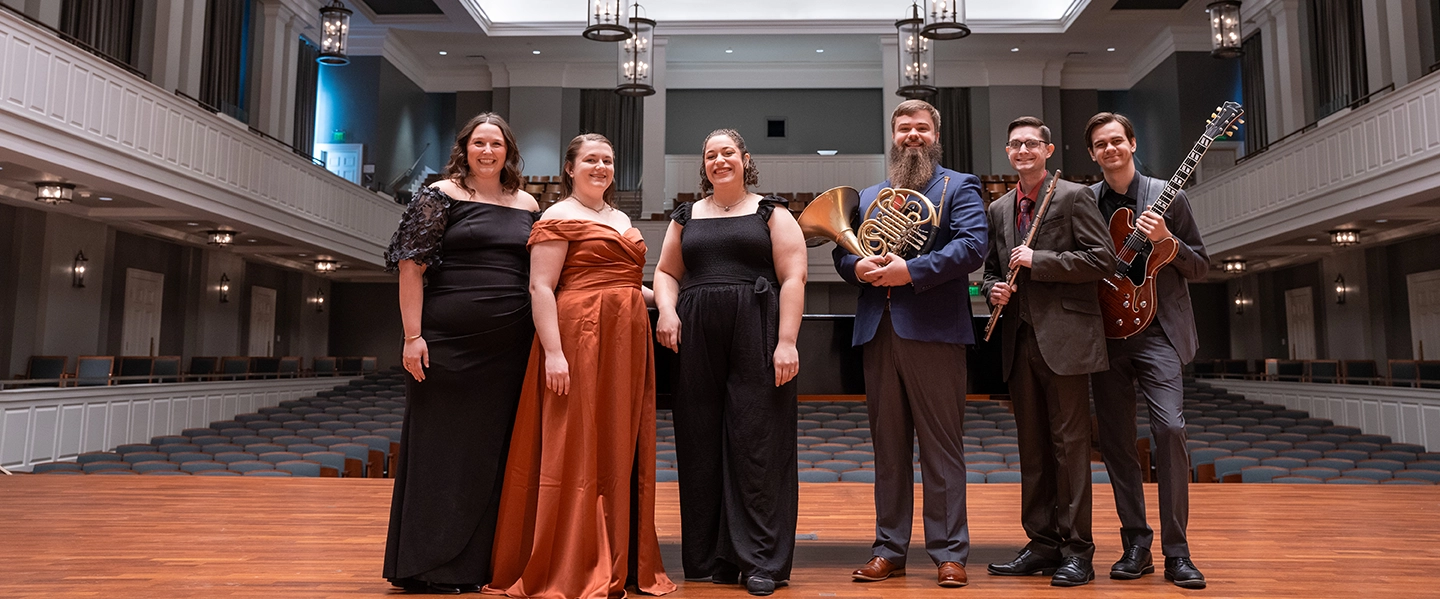 Six musicians stand on stage in an elegant concert hall, dressed formally, with two holding instruments—a French horn and a guitar.