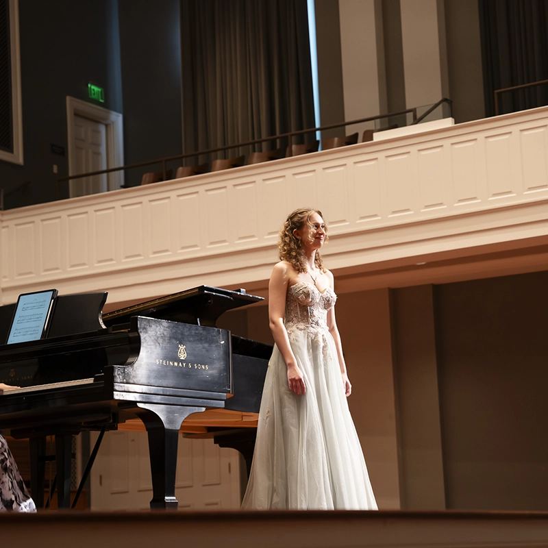 A woman in a formal gown stands on stage beside a Steinway grand piano, smiling toward the audience in an elegant concert hall.