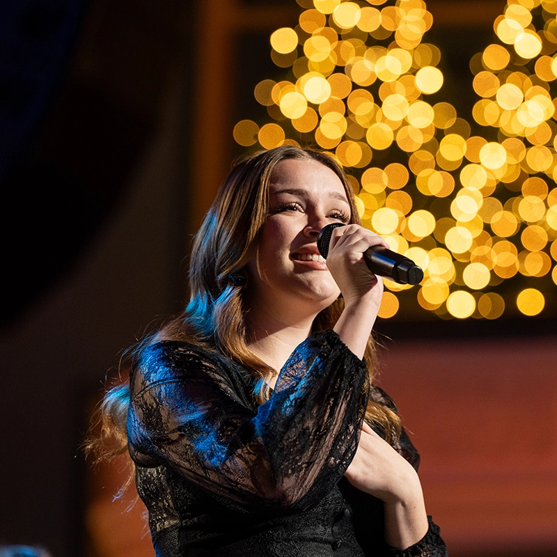 A woman in a black lace top sings into a microphone on stage, with warm golden lights blurred in the background.ge, wearing a dark velvet costume with a white shirt underneath. The background is lit in shades of red, enhancing the dramatic atmosphere of the vocal performance.
