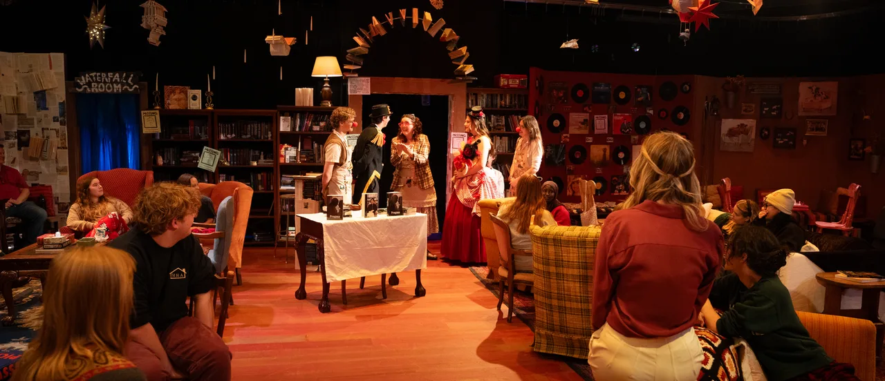 A theatrical stage performance in an intimate theater setting with warm amber lighting. Actors perform on a library-themed set decorated with bookshelves, furniture, and hanging props including lanterns and decorative elements. An audience sits in chairs surrounding the stage on an orange floor. A sign reading "Waterfall Room" is visible in the background.