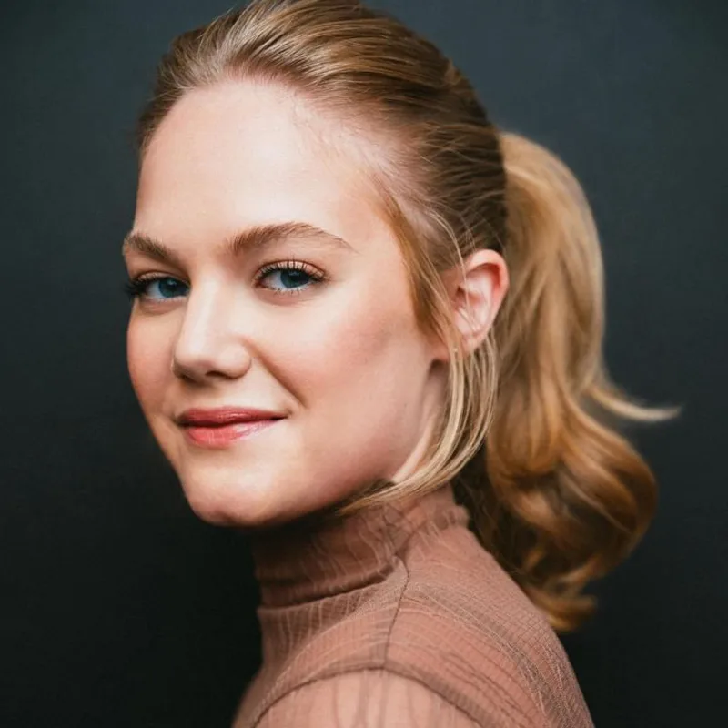 Close-up portrait of a woman with blonde hair in a ponytail, looking over her shoulder with a slight smile against a dark background.