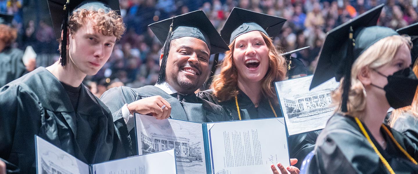 3 students sitting at graduation ceremony in cap and gown as they hold their diplomas and pose for a picture