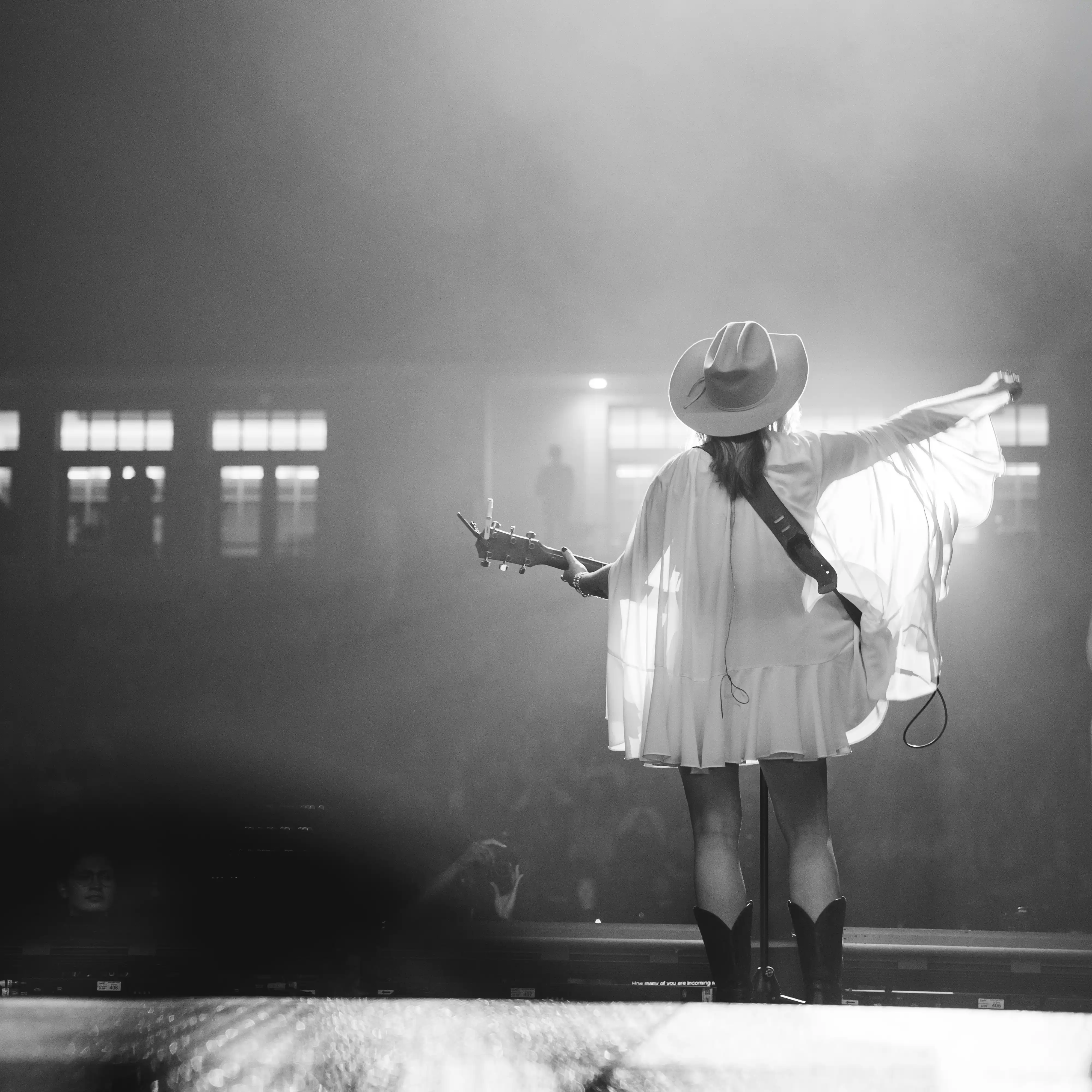 A black-and-white photo of a musician on stage, captured from behind. The performer, wearing a wide-brimmed hat, boots, and a flowing outfit, holds a guitar and raises one arm to the side, illuminated by stage lights in front of a live audience.