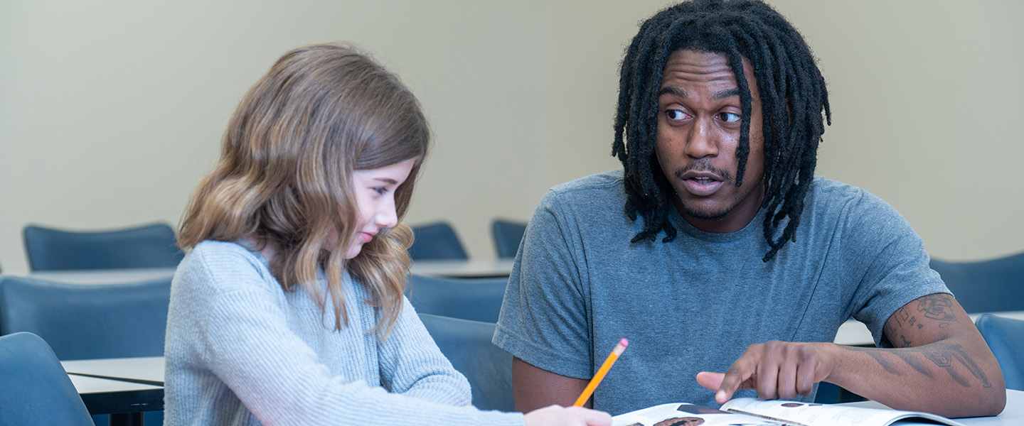 A male teacher sits with a young student and directs her attention to a book that he's reading from.