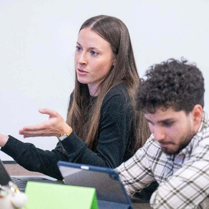 Woman speaking and gesturing during a meeting or lecture, next to a man using a digital tablet.