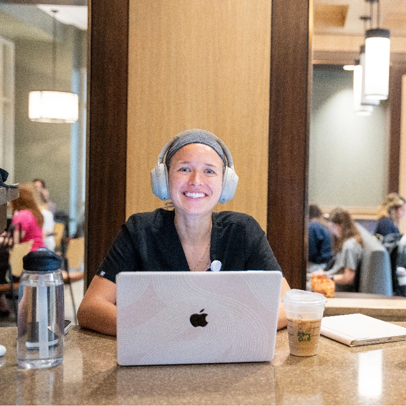 Smiling woman in scrubs with headphones working on MacBook laptop, studying in a busy common area.