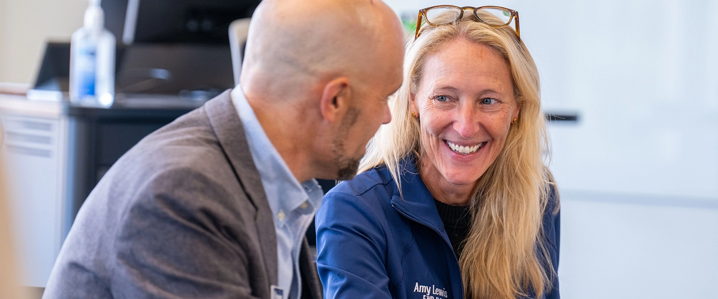 A woman with long blonde hair speaks with a man in a suit while the two are seated at a table.