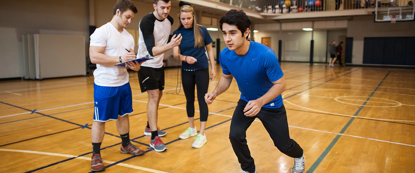 Student in athletic gear preparing to sprint in a gym while classmates observe and take notes for a fitness assessment.