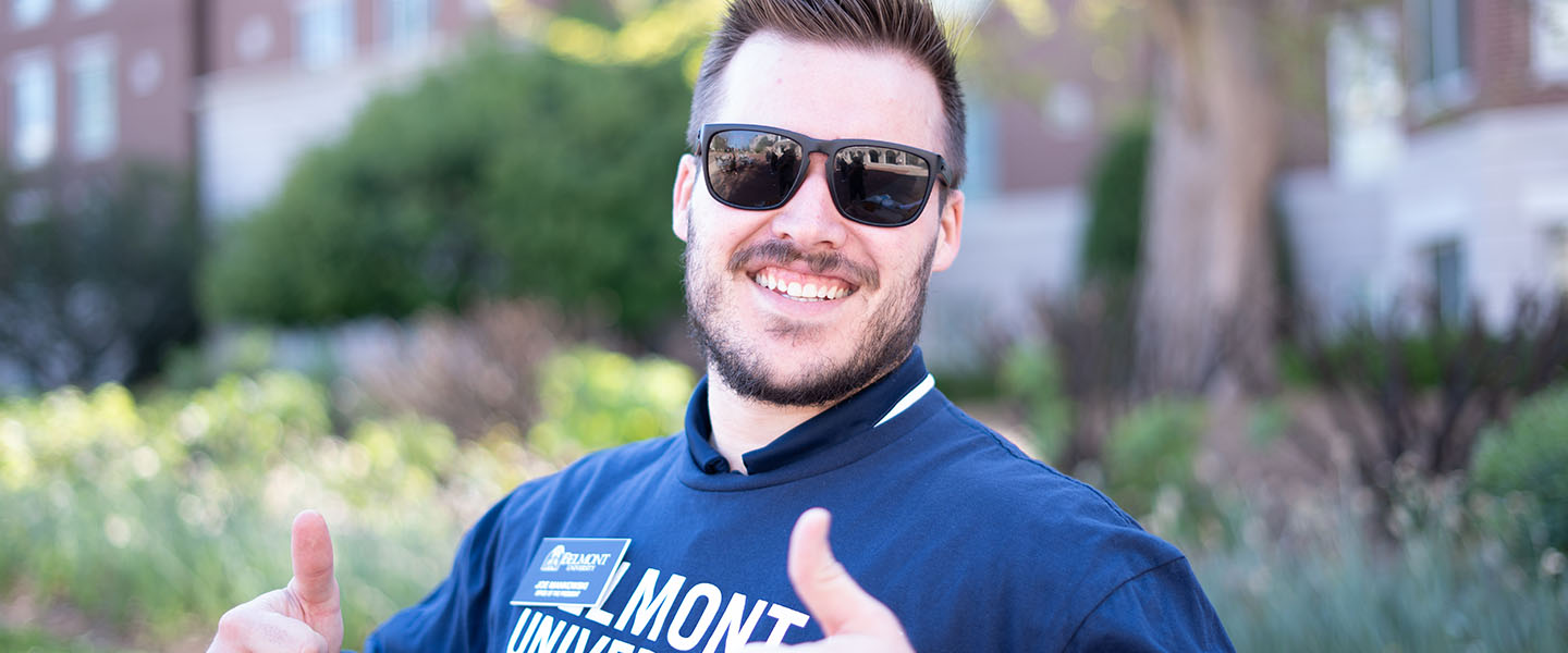 A smiling person wearing sunglasses and a Belmont University shirt giving a thumbs up while standing outdoors on campus.