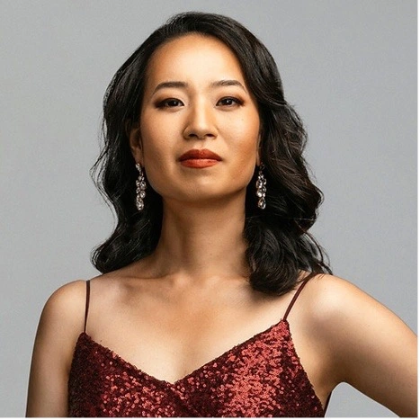 Studio portrait of a woman with wavy dark hair wearing a red sequined spaghetti-strap dress and dangling earrings against a gray background.