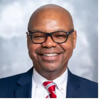 Smiling man in a navy suit wearing black glasses and a red striped tie against a light gray studio background.