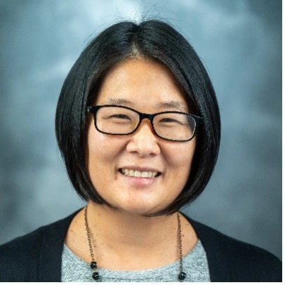 Smiling person with short dark hair and black rectangular glasses, photographed against a gray-blue studio backdrop.