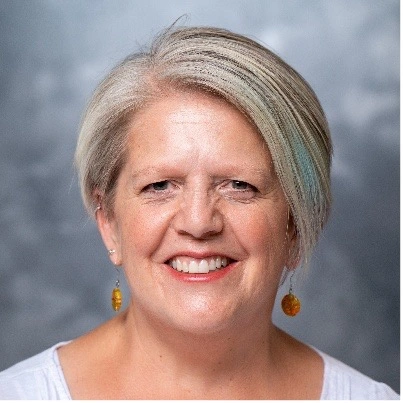 Smiling older woman with short gray hair and amber drop earrings against a gray studio backdrop.