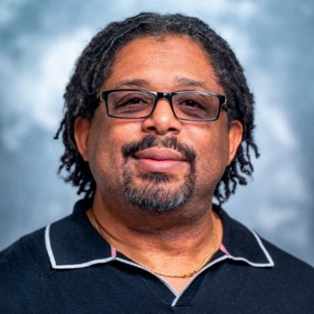 Head-and-shoulders studio portrait of a person with glasses and short dreadlocks wearing a black collared shirt against a mottled blue-gray background.