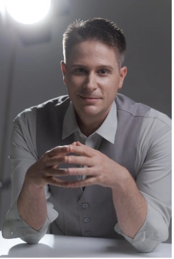 Studio portrait of a man in a light dress shirt leaning forward with hands clasped, with a bright light behind him.