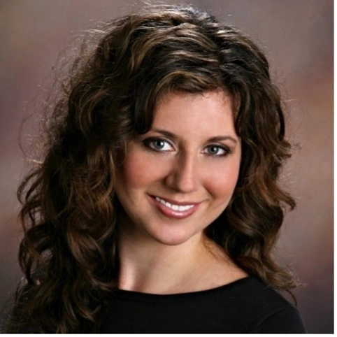Studio headshot of a smiling woman with wavy brown hair against a soft brown background.