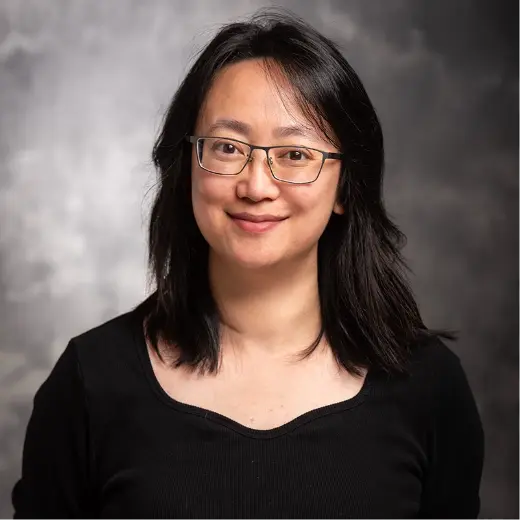 Studio headshot of a smiling woman with shoulder-length dark hair and glasses against a gray background.