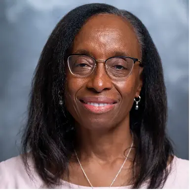 Head-and-shoulders portrait of a smiling woman with glasses and long dark hair against a gray studio background.