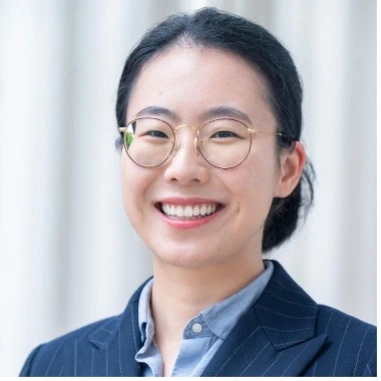 Smiling person wearing round glasses and a dark blazer, photographed in a bright indoor setting.
