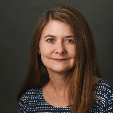 Studio headshot of a woman with long brown hair wearing a patterned navy top against a dark gray background.